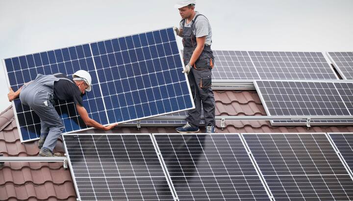 An image of a solar panel installation on the roof of a commercial building in Cumming, GA. The installation team can be seen working on the roof, positioning the panels to capture maximum sunlight. The city skyline can be seen in the background, with clear blue skies and a few clouds. Tools, equipment, and safety gear are scattered on the roof.